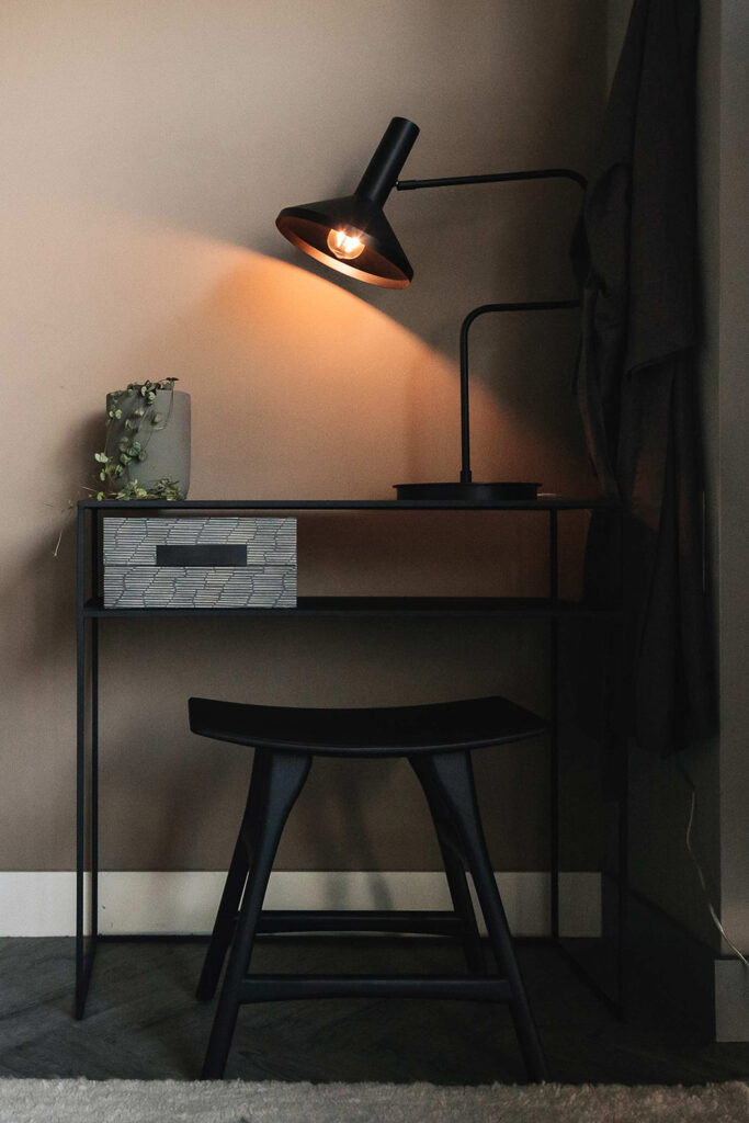 Black metal console table with black stool. Large black desk lamp and small house plant on top.