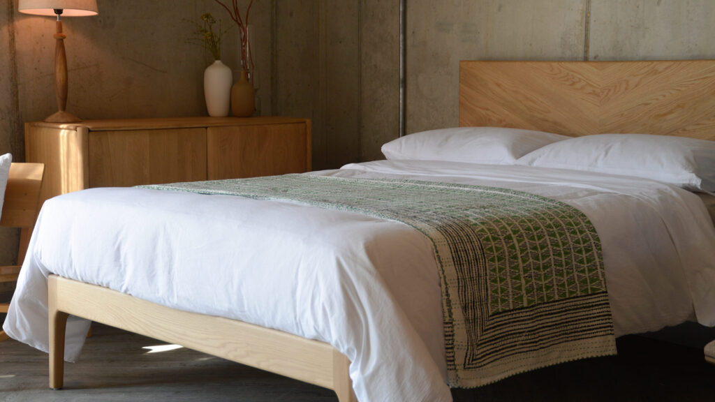 Oak bed frame dressed in white linen with a green triangle pattern bedspread on top. Other oak furniture in room.