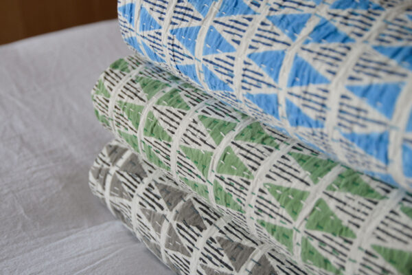 blue green and grey bedspreads with triangle print folded and stacked on a bed with white sheet.
