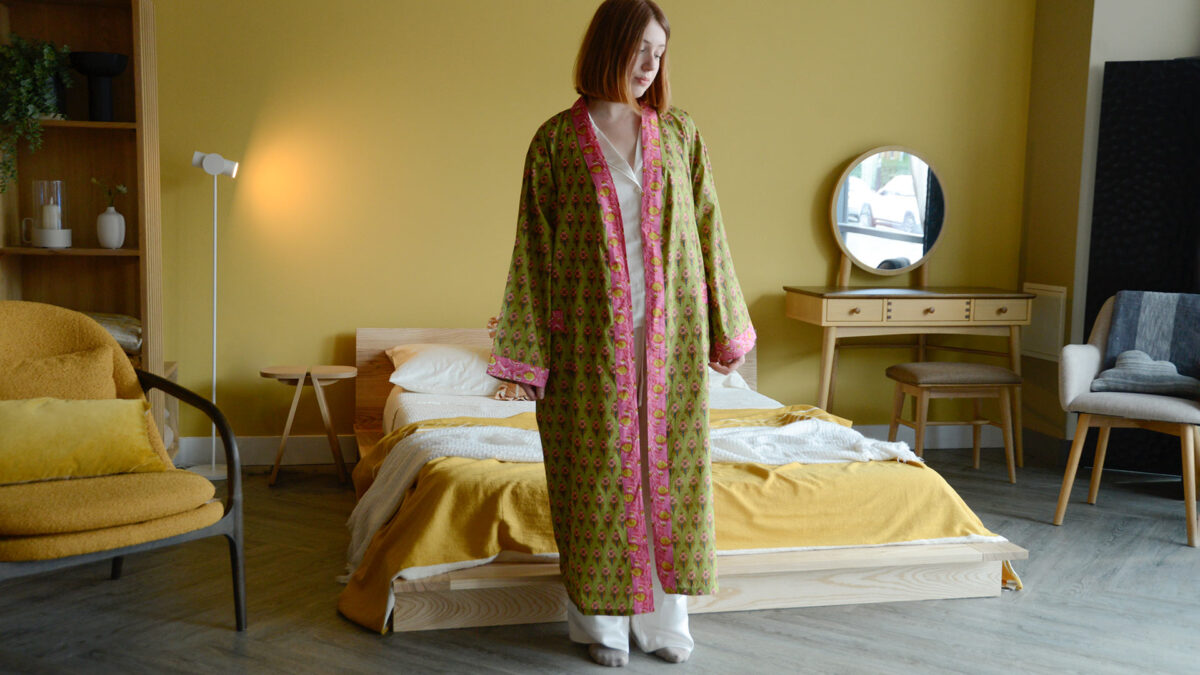 Model stood in room with yellow wall and low, Japanese style bed. Model is wearing satin pyjamas and a green and pink floral robe. Full length image to show size of robe.