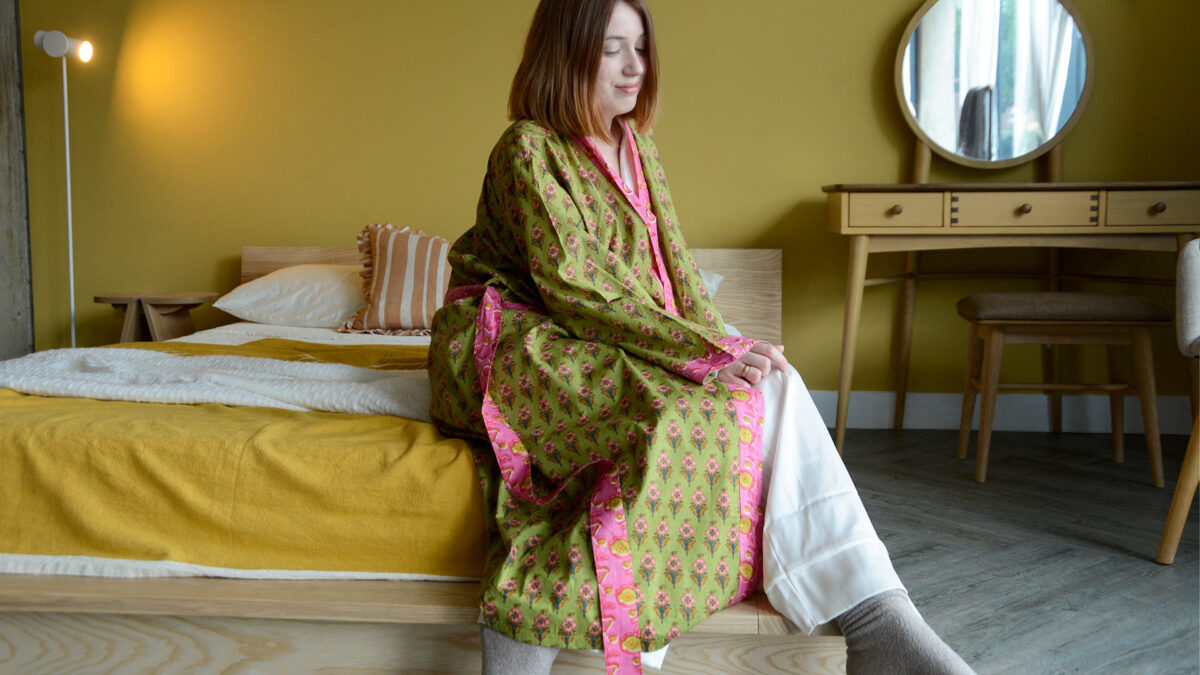 Model sat on corner of bed in room with a yellow wall. Model is wearing a green and pink floral robe with bright pink detailing.