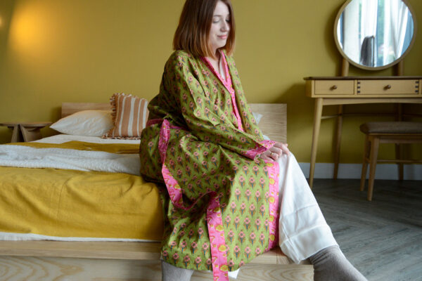 Model sat on corner of bed in room with a yellow wall. Model is wearing a green and pink floral robe with bright pink detailing.