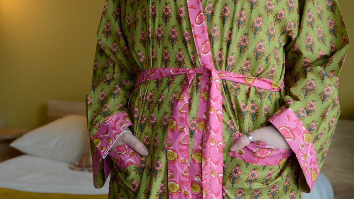 Image showing cropped view of green and pink floral robe on model. Image shows model with hands in pockets of robe, and pink trim found on the edges of the robe, sleeve cuffs and pockets.