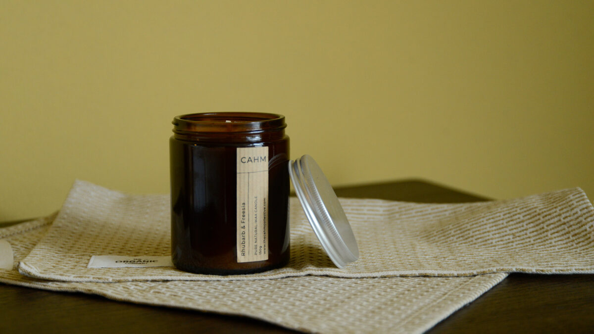 One open Rhubarb and Freesia candle in dark glass jar, lid leaning to the side. Standing on top of a beige cloth on a dark walnut counter top. Yellow wall in the background.