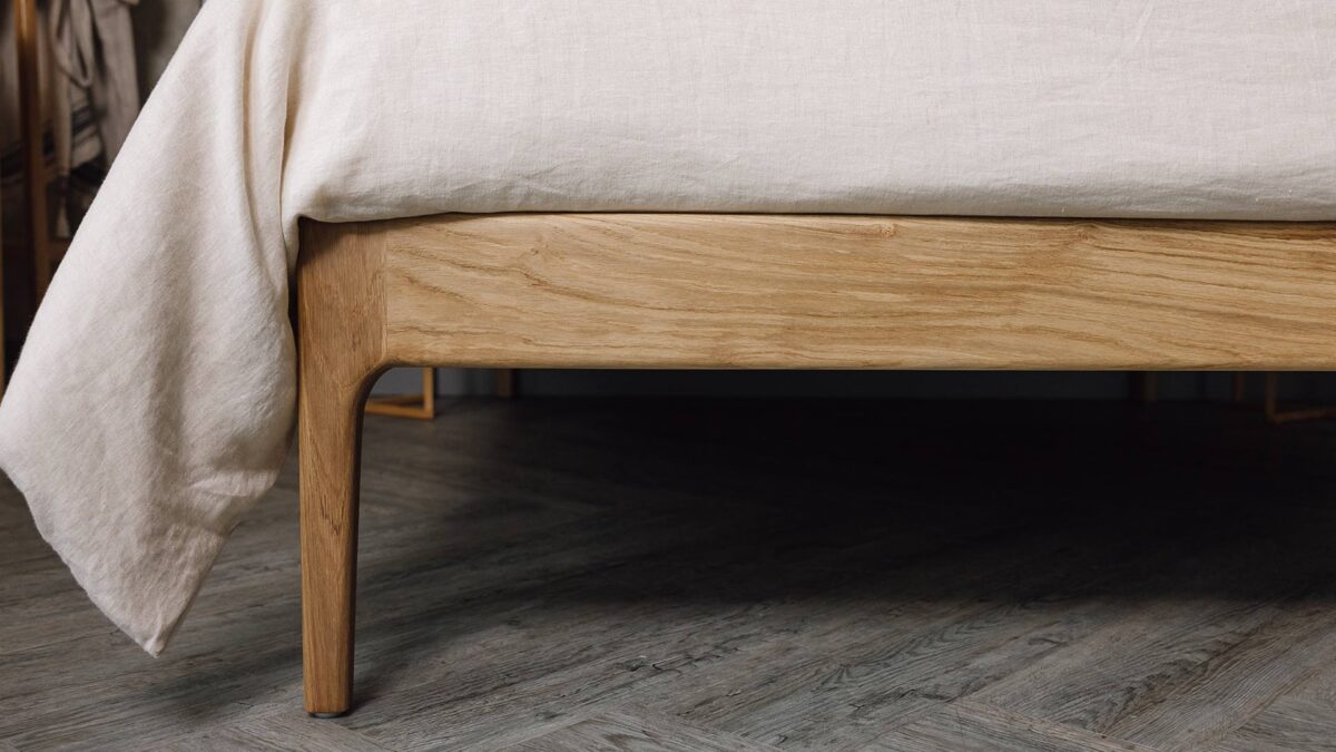 Close up shot of the English oak Camden's foot-rail. The bed is dressed in the organic ecru linen bedding.