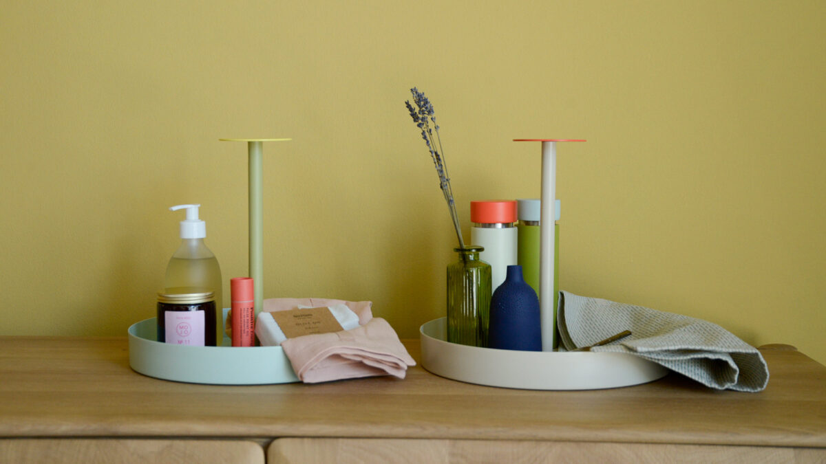 2 Colourful round trays with handles. One light green with yellow topped handle carrying hand soup, a candle and other bathrooms supplies. The other grey with orange handle carrying a vase and flower, a cloth and a colourful salt and pepper grinder.
