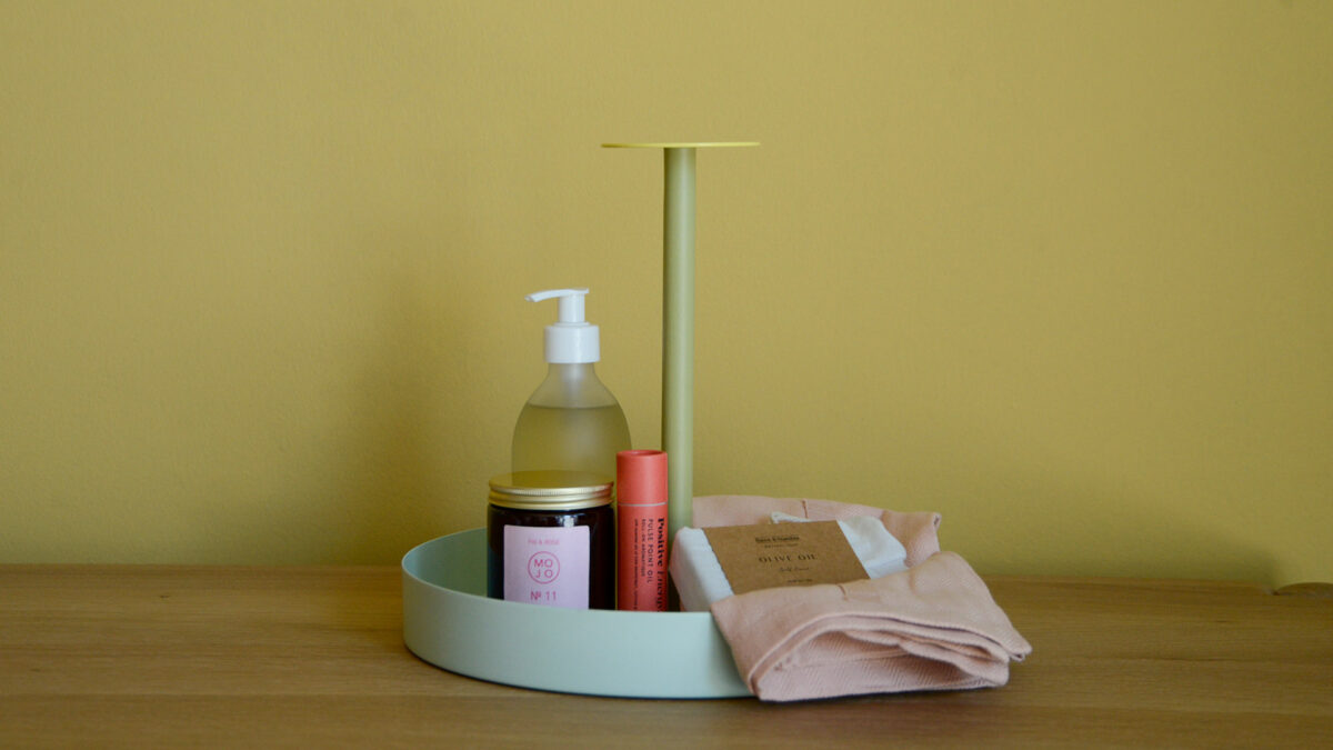 Round metal tray in green sitting on an oak unit in front of a yellow wall. Bathroom supplies on tray.