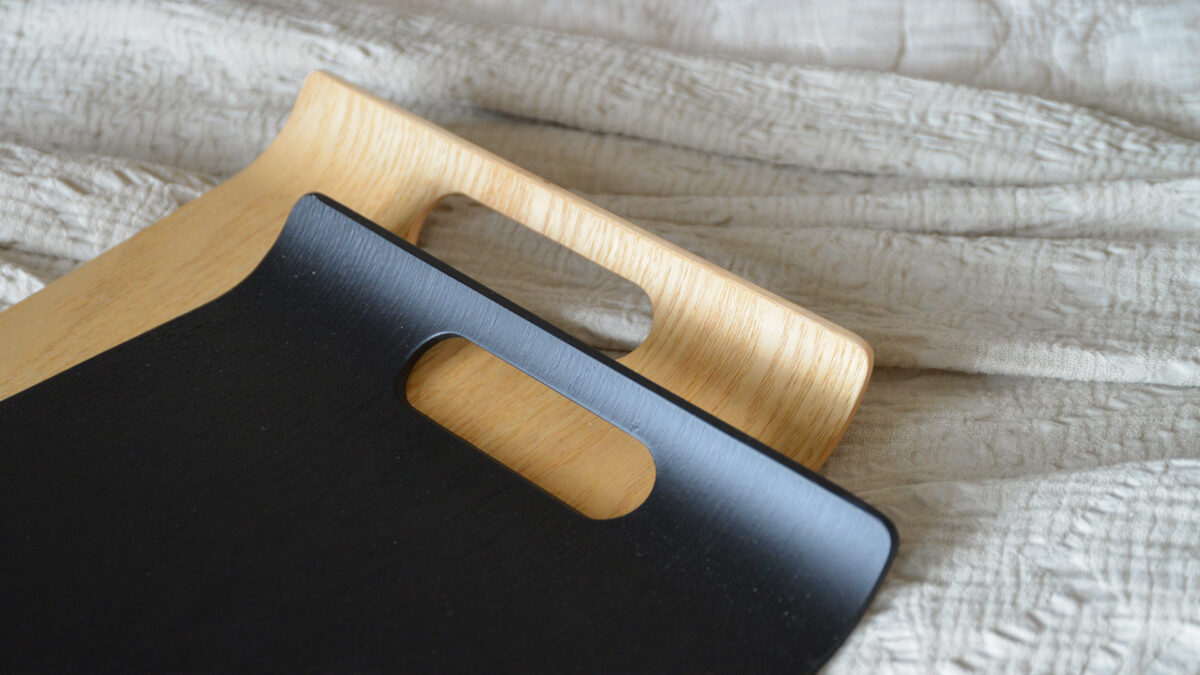 2 curved trays, one black, one oak, laid on a white textured bedspread.