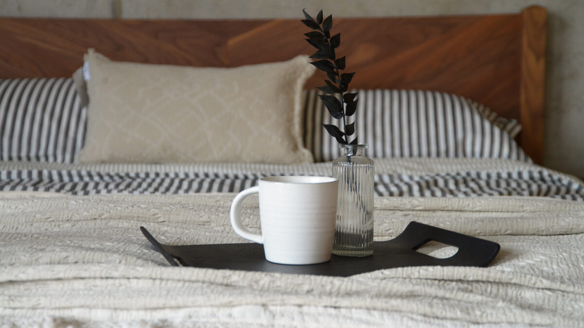 Curved black tray with a white mug and small glass vase laying on a bed.