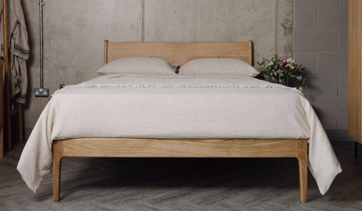 A shot of the English Oak Camden, showing its headboard and focusing on its foot-rail primarily. The bed base is dressed in the organic linen bedding and the floral textured throw in the ecru colour-ways.