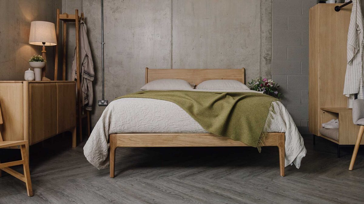 front view of an English Oak Bed Frame with beige linen, a textured bedspread and an olive green merino wool throw. Bedroom scene also shows an oak sideboard, a coat stand, a chair and an open clothes rail.