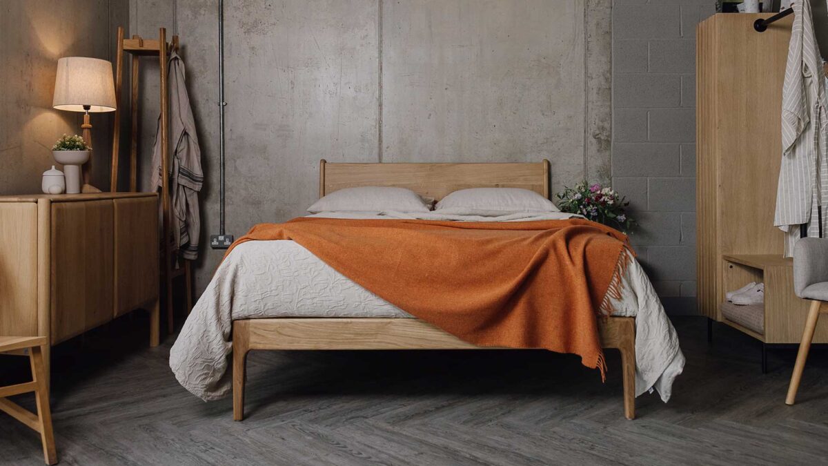 Front view of a bedroom scene with an English oak bed in the centre, surrounded by an oak sideboard, a coat stand with robe hanging on it and an open rail and shelf set. Bed made with ecru linen and a beige textured bedspread, with a burnt orange lambswool throw draped over the top.