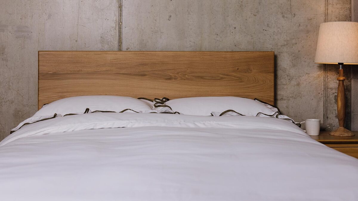 A zoomed in image of the English oak Chiswick, showing it dressed in the white bedding with a green ruffle-edge detailing. The image shows only the bedding and the Chiswick's headboard panel against a concrete wall.