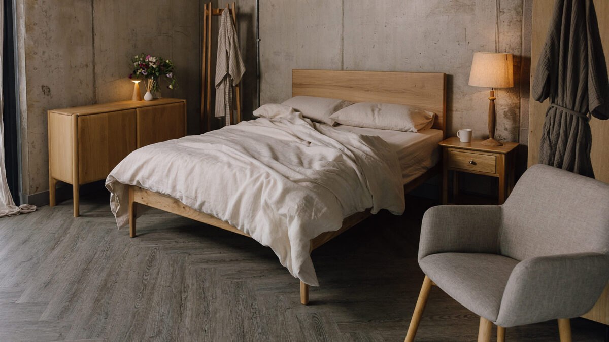 3/4 angle image of oak bed with large wooden headboard. Bed dressed in rumpled linen, other oak furniture in the room scene.