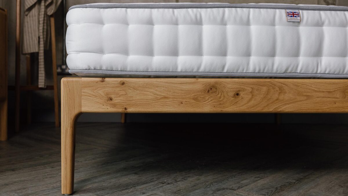 A close up shot of part of the English oak Chiswick's foot-rail/leg. The foot-rail has several knots in it. The top part of the image shows that the bed has a bare mattress on it.