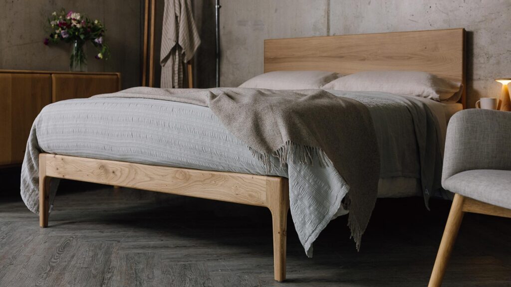 A low angled shot of the English oak Chiswick, focusing on the foot-rail section of the bed. The bed is dressed in the organic linen ecru bedding, a silver grey embroidered bedspread and a beige pure wool throw.