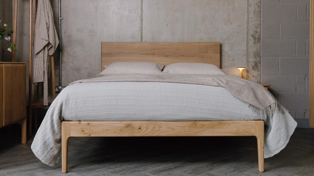 A forward facing shot of the English oak Chiswick, showing its foot-rail/legs and the headboard panel. The bed is dressed in the organic linen ecru bedding, a silver-grey embroidered bedspread and a beige pure wool throw.