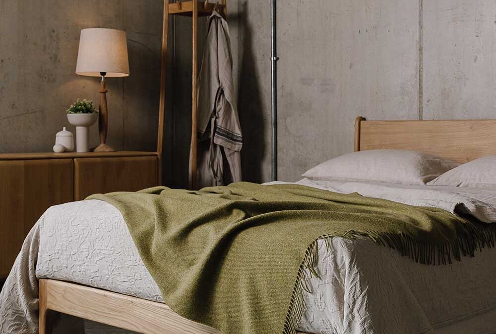 3/4 view of a handmade English Oak Bed Frame with beige linen and a Green lambswool throw. Oak sideboard and coat stand in the background.
