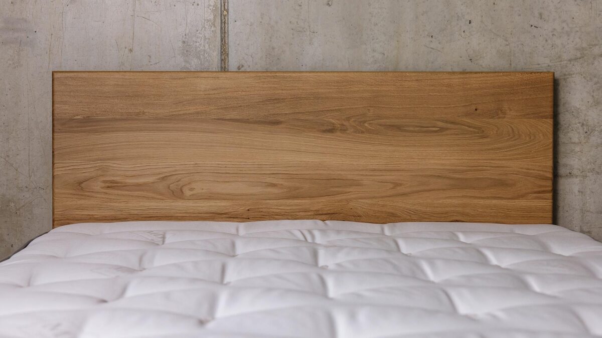A close up shot of the English oak Chiswick's headboard panel against a concrete wall. There is a bare mattress on the bed base.