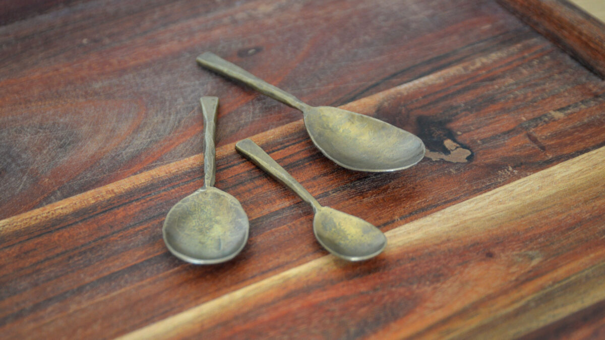 Forged iron spoons with a gold/brass colour in three sizes. The spoons are sat on a meh wood tray, finished with a dark reddish brown stain.