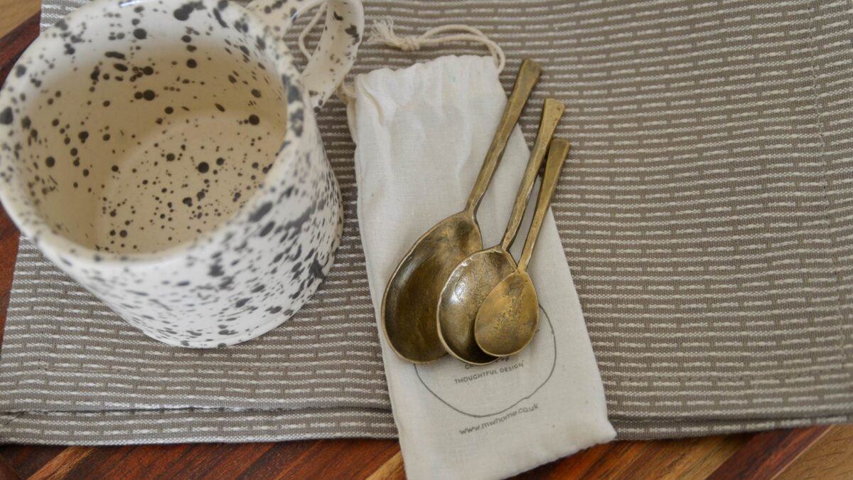 Forged iron spoons sat on the bag that they come packaged in, to show how they will arrive to you. Pictured also is a grey and white mug to give a sense of setting.