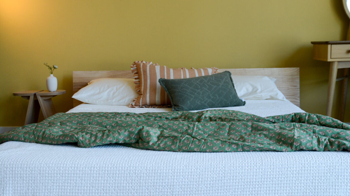 Image shows a green floral quilted throw on a low bed with one green and one yellow cushion. The bed is dressed with a pure white bedspread and sat against a yellow wall.
