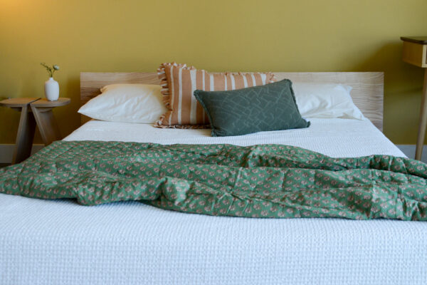 Image shows a green floral quilted throw on a low bed with one green and one yellow cushion. The bed is dressed with a pure white bedspread and sat against a yellow wall.