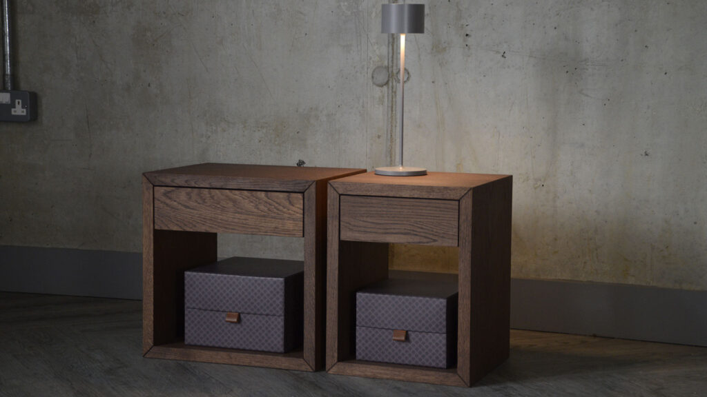 Image shows two bedside tables with a dark brown, walnut-look finish. The tables feature an upper drawer and lower shelf, which house some purple storage boxes.