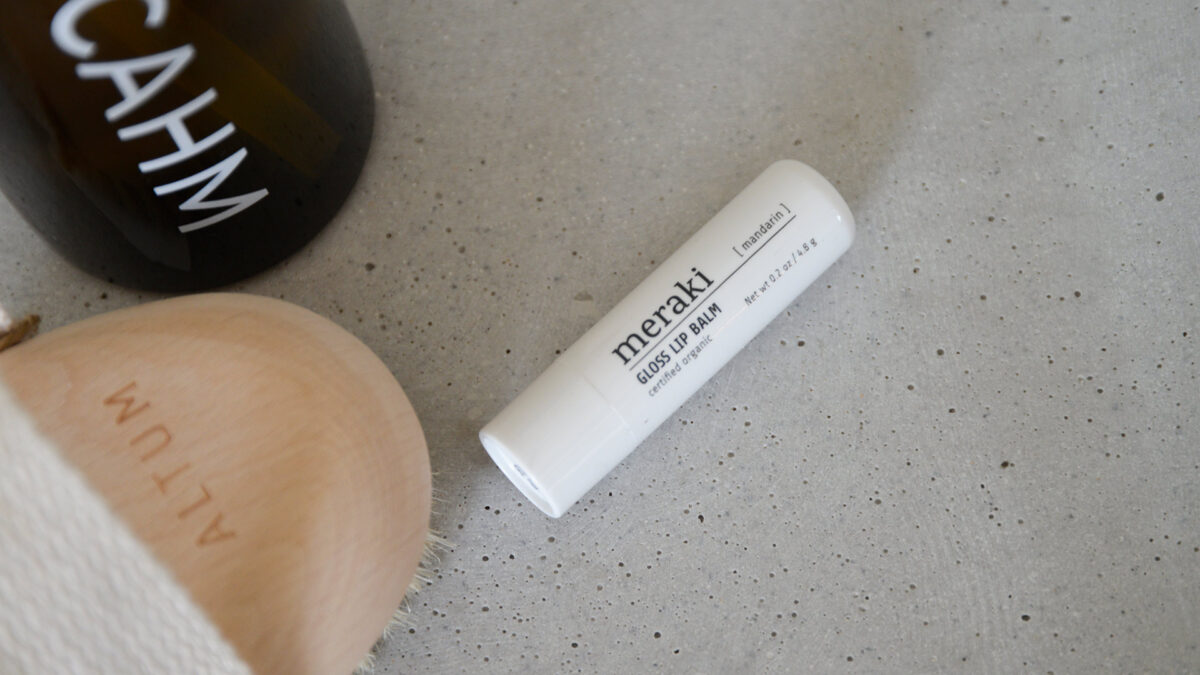 A closeup image of a gloss lip balm from Meraki. Other toiletries can be seen in the background to give a sense of setting.