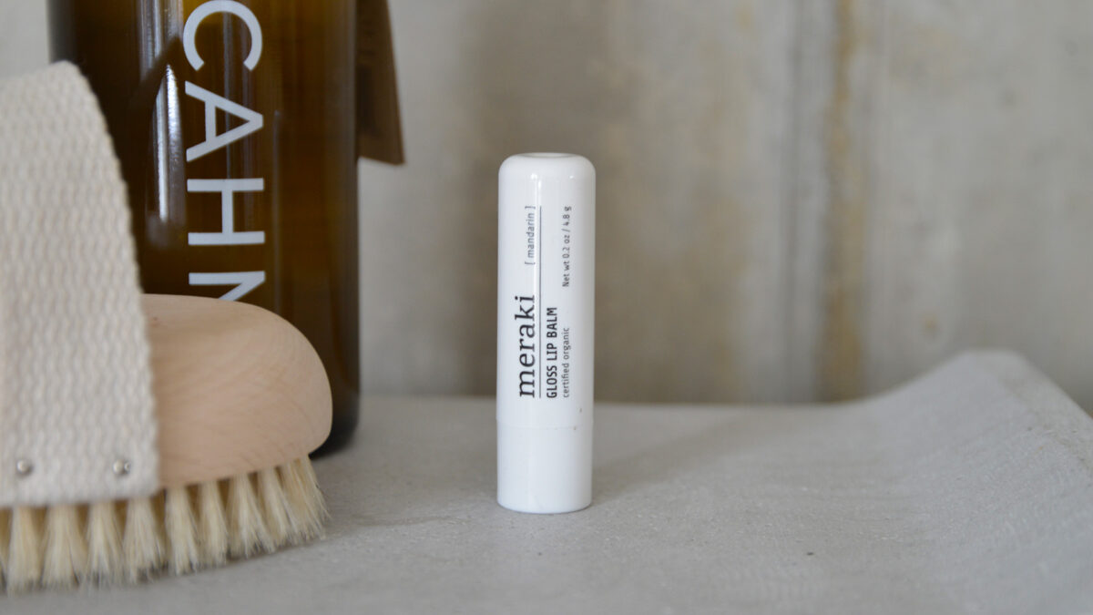 A closeup image of a gloss lip balm from Meraki. Other toiletries can be seen in the background to give a sense of setting.