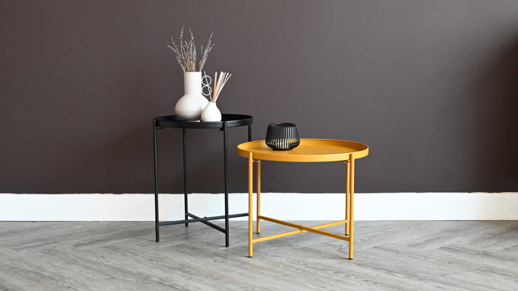 2 round metal tables. One taller, but smaller in black, the other shorter and wider in mustard yellow. Vases and candle holders on top.