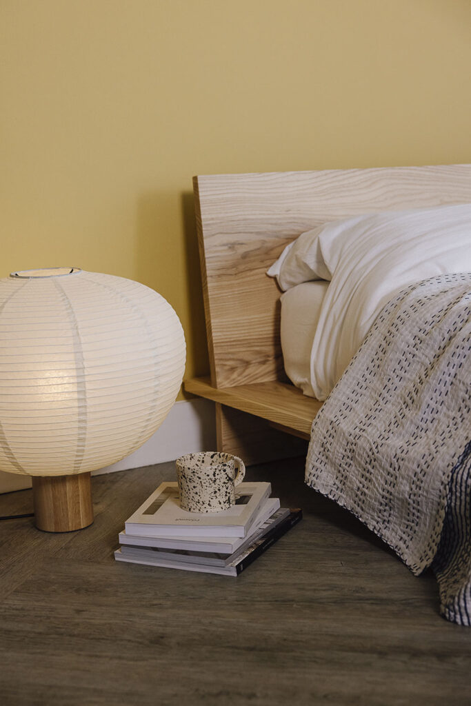 Portrait image of the top corner of a modern loft bed in ash wood with a white duvet cover and pillow cases and a blue and white bedspread with stitching detail. Japanese inspired lamp and stack of books to the side.