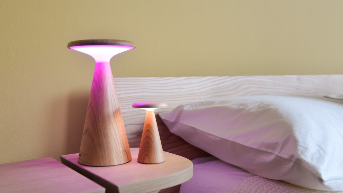 2 Oak table lamps, one large and one small, switched on, sitting on a bedside table next to a bed.