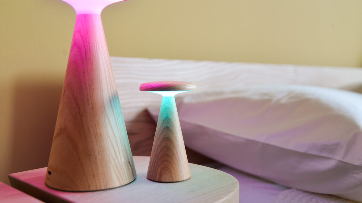 2 Oak table lamps, one large and one small, switched on, sitting on a bedside table next to a bed. Large light lit in purple, small in green.