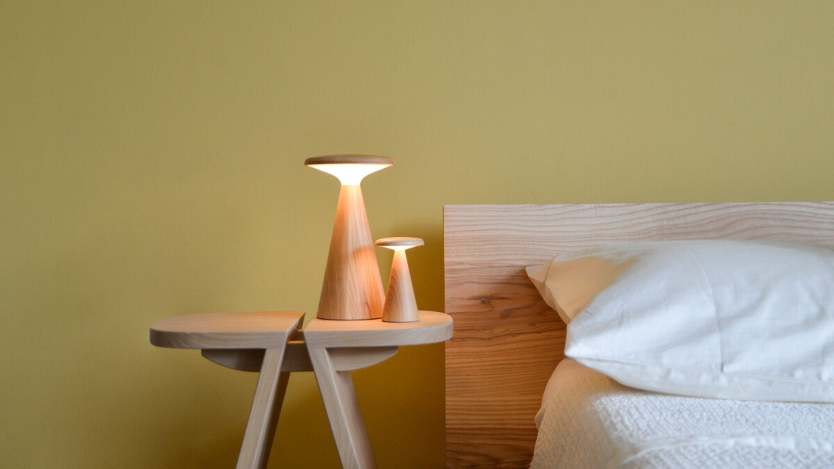 2 Oak table lamps, one large and one small, switched on, sitting on a bedside table next to a bed.