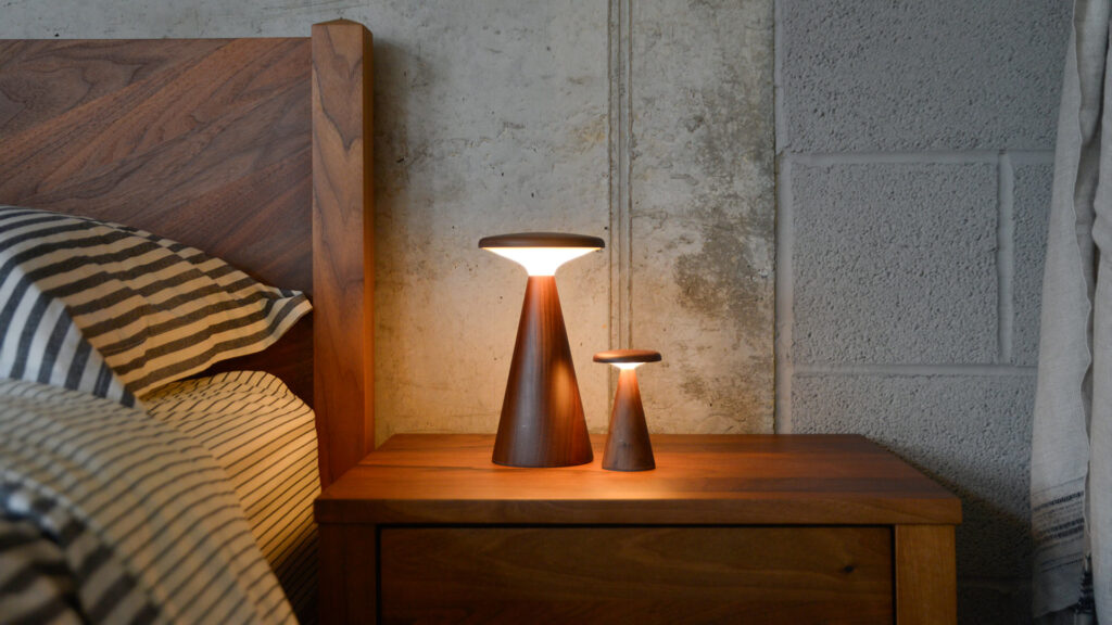 2 walnut table lamps in the same design, but one large and one small. Lit with white light, sitting on a bedside table next to a bed.