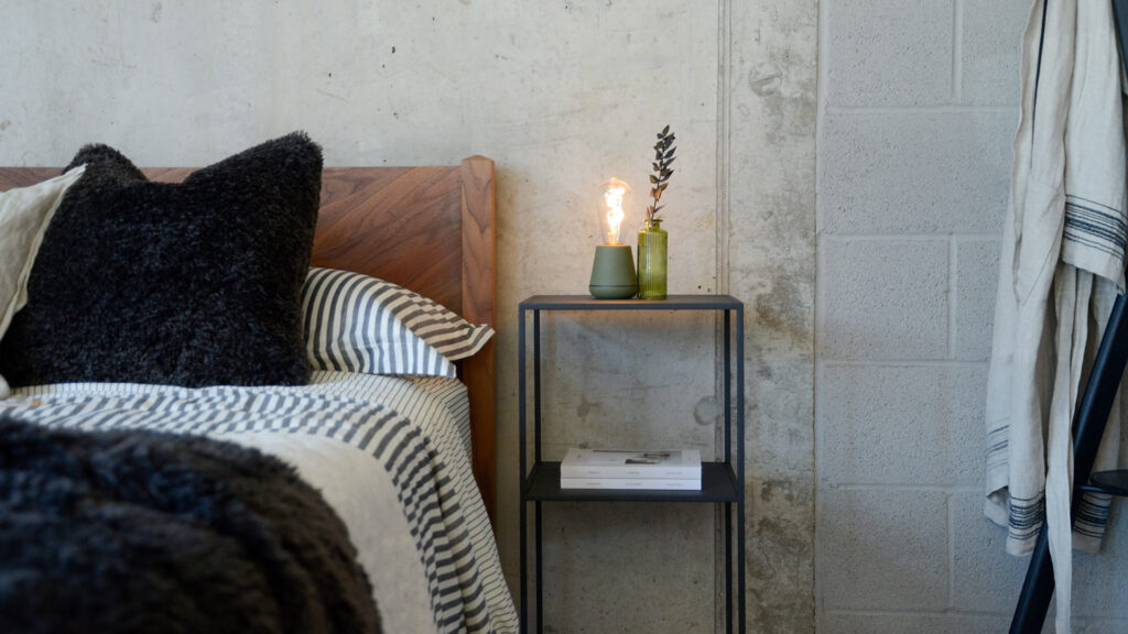Zoomed out image of the tall Fera powder coated metal table in dark charcoal grey, stood next to the Dalston bed in walnut.