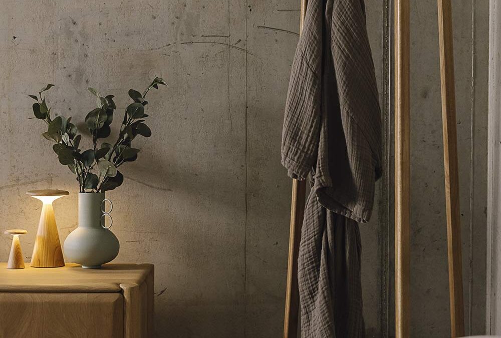 Portrait image showing the Tower Coat stand with a clay coloured organic cotton robe hanging from it. To the left, you can also see part of a solid oak unit from Ethnicraft, with some small accessories - including a lamp and vase - sat atop it. To the bottom of the image, you can see a bed base, but this is not the main focus of the image.