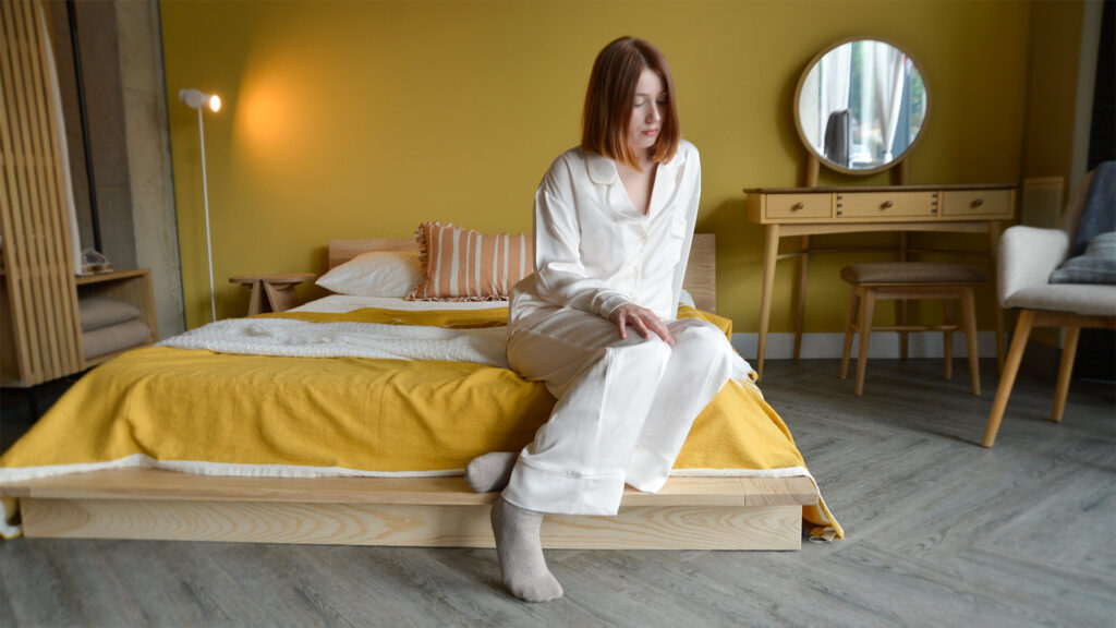 Woman wearing white satin Pyjamas sat on a bed with yellow bed linen.