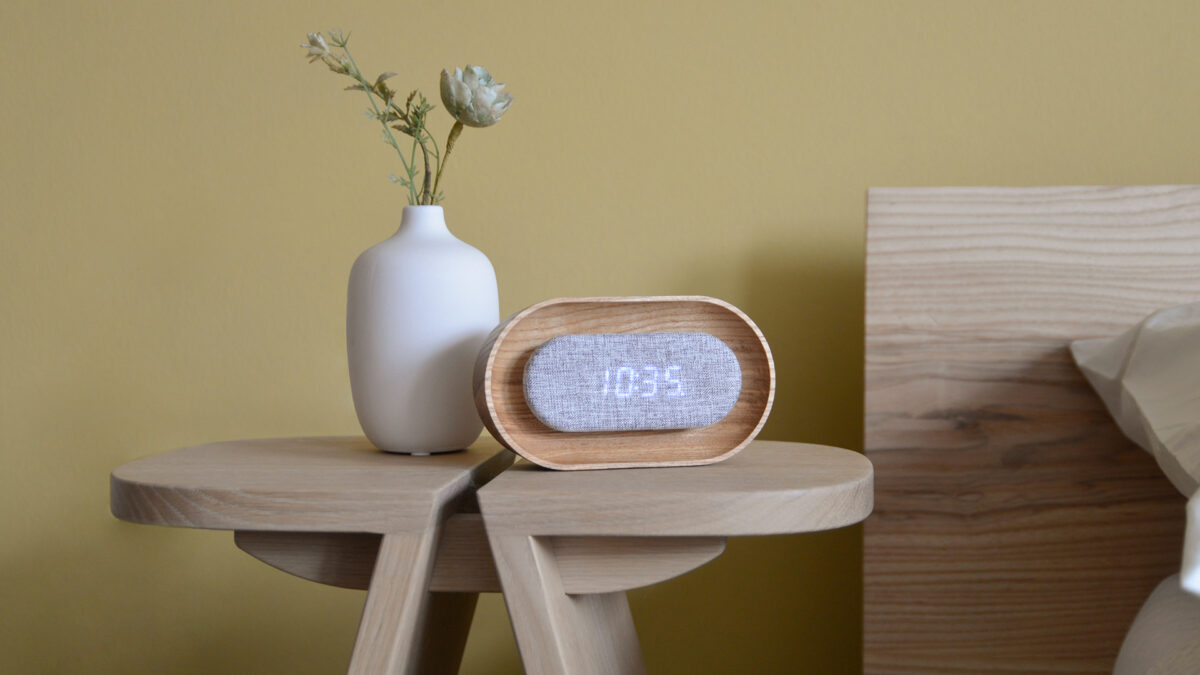 Oak alarm clock with digital display, photographed on table to the side of a bed.