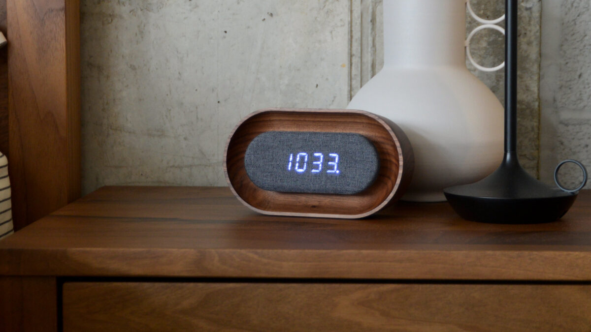 Walnut alarm clock with central digital display sitting on bedside table with vase and lamp.