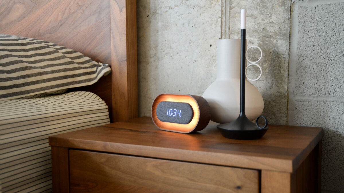 wooden alarm clock with digital display photographed on a bedside table with a vase and lamp.