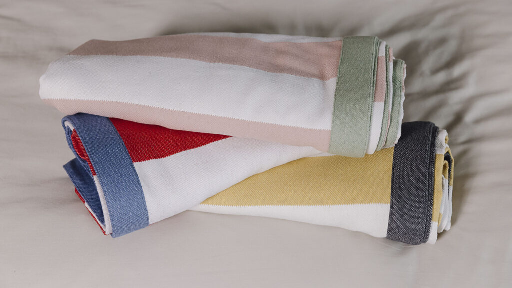 Three striped throws with contrasting border rolled on bed. From bottom to top, yellow and white with grey border, white and red with blue border, pink and white with sage green border.