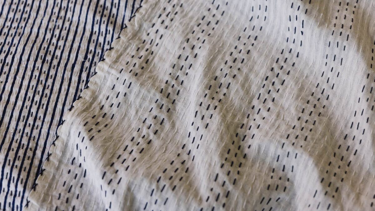 Close up of a deep blue kantha style bedspread, folded over so that both the front with a blue stripe and back with a blue stitching detail are visible.