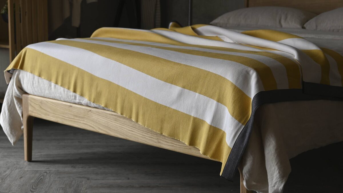 Striped yellow and white throw with grew border draped across a bed with ecru linen.