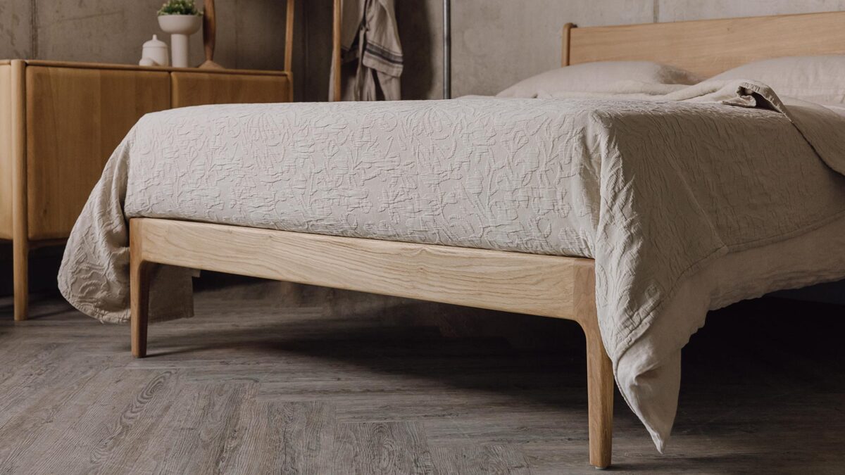 An image of the end of the English oak Camden bed base, focusing on the foot-rail and the floral textured bedspread in the ecru colour-way.