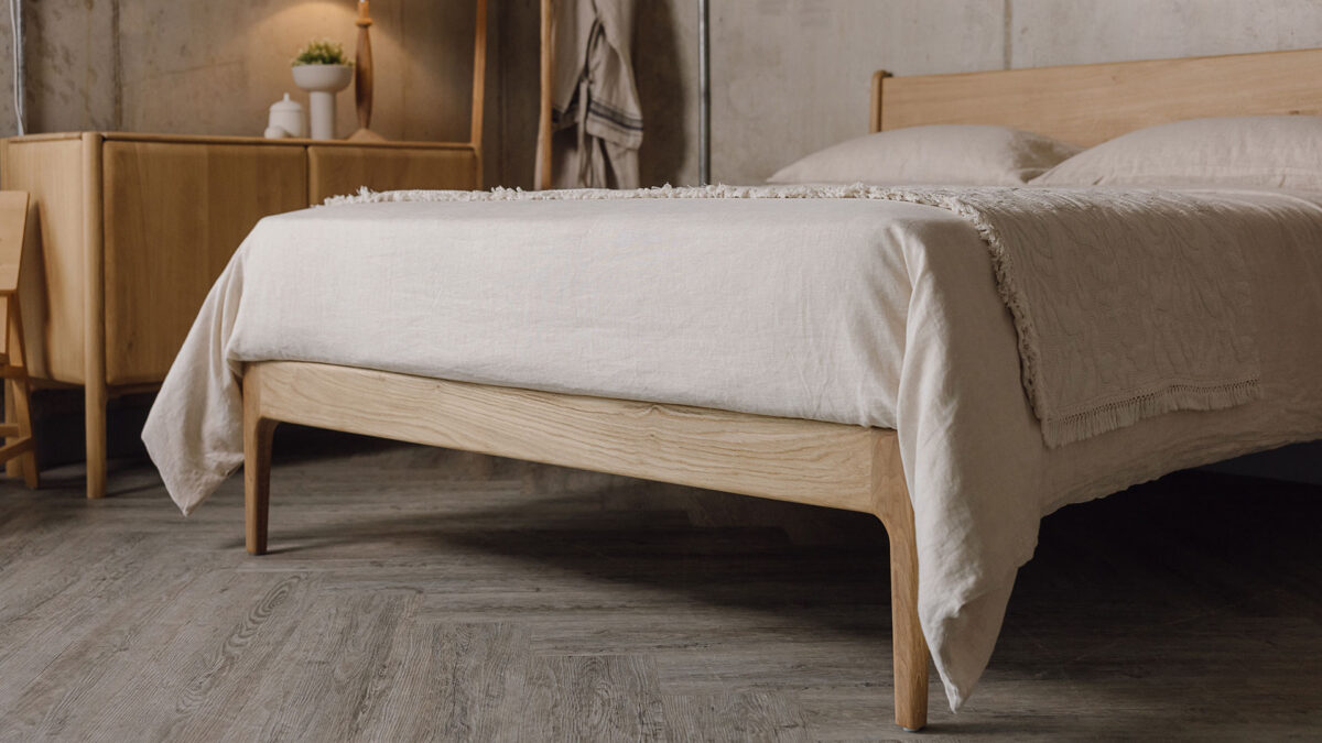 Close up angled shot of the end of the oak Camden bed base, showing the curved detail of its legs. The bed base is dressed in ecru linen bedding with a textured ecru throw with fringe detail laid on top.