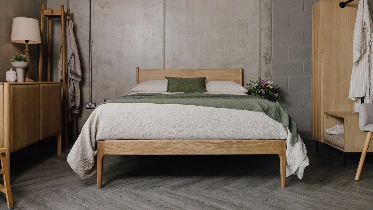 Front on shot of an English Oak Camden bed in a bedroom scene. Bed covered in beige linen and pillow cases, with a textured beige throw, topped with a textured olive green throw and a small olive green cushion.