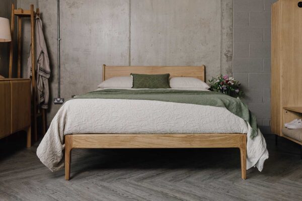 Front on shot of an English Oak Camden bed in a bedroom scene. Bed covered in beige linen and pillow cases, with a textured beige throw, topped with a textured olive green throw and a small olive green cushion.