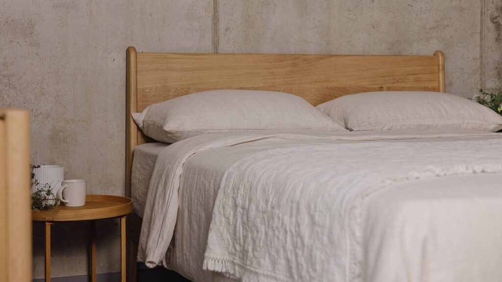 Zoomed out shot of the English oak Camden's headboard panel. The bed is dressed in the organic linen bedding and the floral textured throw in the ecru colour-ways. There is a round-topped oak bedside table next to the bed with a white plant pot and a beige coffee mug on top of it.